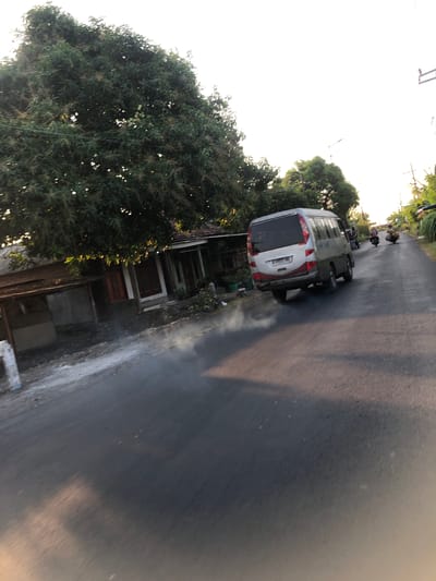 The image captures a street scene in Glagah Arum, Indonesia, during what appears to be late afternoon or early morning, given the lighting. A silver minivan is traveling on a paved road, emitting a plume of white exhaust from its rear. To the left of the road, there are houses with red-trimmed windows and a dense tree with dark green foliage. The ground on the left side of the road appears to be dirt with some white debris. In the distance, two motorcycles are also visible on the road, with riders on them. The sky is bright and hazy, suggesting either sunrise or sunset. The overall impression is of a quiet, possibly rural or suburban, residential area with everyday traffic.