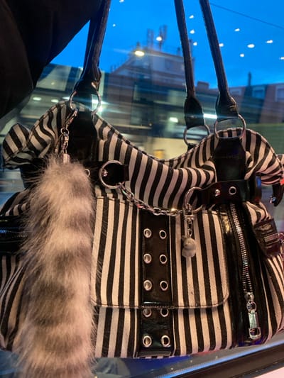A black and white striped handbag is prominently featured in the foreground, positioned as if hanging or placed against a reflective surface. The bag exhibits vertical black and white stripes, interspersed with elements of black patent leather, including the two shoulder straps, a central vertical panel with six silver-toned eyelets, and additional accent straps with buckles and eyelets. A silver-toned metal chain runs across the front of the bag, connecting two D-rings. Attached to the left side of the bag via a small silver-toned keyring is a fluffy, grey-brown fur or faux fur accessory. On the right side, another small silver-toned keyring holds a light grey, possibly fabric or plastic, charm resembling a small animal or figure. A silver-toned zipper with a prominent pull tab is visible on the right side of the bag, extending vertically along a patent leather trim.

The background is a reflection seen through a glass pane, appearing distinctly blue, indicative of a twilight or night sky. Reflected in the glass is a multi-story building with a light-colored facade, featuring darker rooflines and what appears to be a telecommunications tower or similar antenna structure on its rooftop. Several yellow lights, possibly streetlights or building illuminations, are visible as distinct points of light and reflections within the blue background. The reflections also show faint, blurred horizontal lines, suggesting movement or a distant environment. The scene depicts an object likely viewed from inside a vehicle or building, looking out onto a street or urban landscape in Enghien-les-Bains, France, during low-light conditions. No people are visible.