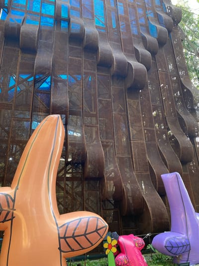 The image shows an outdoor scene with a large, abstract metal sculpture in the background, made of perforated metal panels that undulate in a wave-like pattern. Behind the metal structure, a bright blue material is visible, possibly a ceiling or translucent panels. In the foreground, there are several large, colorful inflatable objects. On the left is a large, orange, star-shaped inflatable with black lines resembling veins on its "arms." To the right of the orange inflatable, a bright purple, finger-like inflatable is positioned. Between these two, a pink inflatable object is visible, with a yellow and red flower decoration in front of it. The ground appears to be covered in lush green foliage and some smaller plants. The overall impression is of an art installation in a public outdoor space. The metadata indicates the location as Ciwaruga, Indonesia, suggesting this is likely an art piece displayed in a park or cultural area in that city.