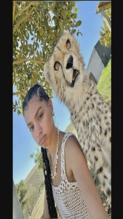 A young woman with braided hair and a shell necklace poses for a selfie in a sunny outdoor setting. She wears a white crochet top. Behind her, a taxidermied cheetah head and neck is propped up, giving the impression of the animal standing behind her. The background includes trees, a grassy area, and a building. The sky is visible above the foliage.