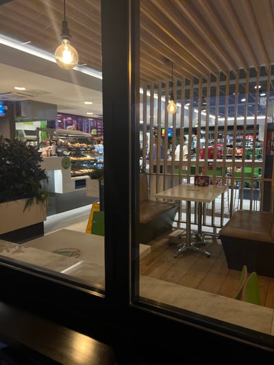 The image shows an interior view of a McDonald's restaurant in Šempeter pri Gorici, Slovenia. The scene is viewed from outside, through a window, at night. Inside, a seating area with light brown booth seats and a light-colored square table with a chrome base is visible. The table has a small, colorful box on it. Beyond the seating area, a serving counter with food displays and menus is visible. Several people are working behind the counter, and customers appear to be present. The interior features a modern design with wooden slatted ceiling and partitions. A hanging pendant light with a visible bulb illuminates the foreground seating area. The floor in the seating area is wooden planking. In the background, shelves stocked with various products are visible.