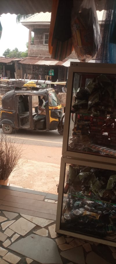 The scene depicts a street view in Umuoji, Nigeria, observed from the interior of an apparent retail establishment.
In the immediate foreground, to the left, a bundle of dried, light brown plant stalks or reeds stands vertically. The floor consists of irregularly shaped, light-colored flagstone or tile pieces set in dark grout. To the right, a white-framed display case with clear glass shelves is filled with numerous individually packaged snack items, primarily in clear plastic wrappers. Overhead, suspended from a visible corrugated metal awning or ceiling, a clear plastic bag containing packaged "Rose Biscuits" hangs alongside an orange fabric item.
In the midground, an unpaved or dirt road, reddish-brown in color, extends across the frame. A three-wheeled motorized rickshaw, commonly known as a Keke Napep, is stopped on this road, oriented with its front facing left. The vehicle is predominantly black with yellow accents. A cargo rack on its roof carries several long, light-colored poles or pipes and a yellow object, possibly a tarp or container. A male individual, wearing a black short-sleeved shirt and light-colored trousers or a wrap, occupies the driver's seat. His left arm is visible near the handlebars, and a blue or green coiled item hangs near his right side.
In the background, behind the rickshaw, several structures are visible. A two-story building with a light-colored corrugated metal roof stands prominently, featuring an open-air balcony and a rectangular window on its upper level. Below this, and to the left of the rickshaw, are smaller single-story structures with rusty corrugated metal roofs, possibly stalls or shops, displaying various hanging items, including green packaged products. Trees and vegetation are visible further in the background, beneath a bright sky.