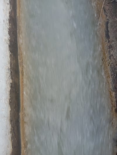 The image shows a close-up, blurred view of a narrow channel of water flowing over a rocky, earthen bank. On the left side, there is a light-colored, rough surface that appears to be stone or concrete, with a dark, irregular line separating it from the water. The water itself is a pale, milky gray and appears to be in motion, creating streaks and blurs. The right bank of the channel is rough and textured, with shades of brown and dark gray, and a thin, dry twig is visible lying on it. The overall impression is of a man-made or natural water channel, possibly in a rural or undeveloped area. The specific location within Kızıl-Bel, Kyrgyzstan, cannot be determined from the image alone.
