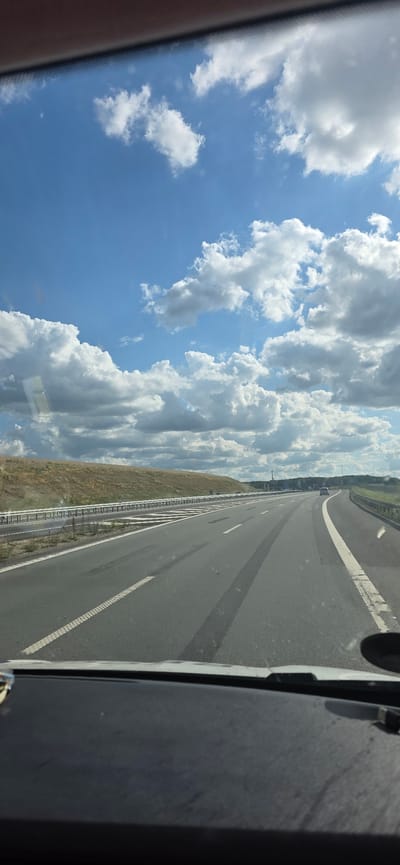 The image is a vertical shot from inside a vehicle looking out at a highway and the sky. The sky is a vibrant blue with scattered white, fluffy clouds. The highway stretches into the distance, curving slightly to the right. It is a multi-lane road with clear white markings. On the left side of the highway, there is a grassy verge and a metal guardrail. A few trees can be seen in the far distance on the left, indicating a rural or semi-rural setting. The overall impression is of a sunny day, likely mid-morning or afternoon given the bright light and clear blue sky. The perspective suggests the vehicle is in motion, traveling along the highway. There is no visible text in the image.