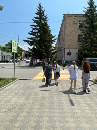 The image captures a street scene in Spassk-Dal'nij, Russia, bathed in bright sunlight, suggesting a warm, clear day. Several people are walking on a sidewalk paved with diamond-shaped tiles. In the foreground, a woman in a dark patterned dress and a yellow bag walks away from the camera, crossing a crosswalk. Behind her, a younger woman with red hair in a braid wears a white shirt and shorts. Further back, another person with long red hair walks in grey athletic pants and a black top. A man in a blue shirt and brown shorts is also visible near the crosswalk.
The setting includes a building with pale beige walls and windows, partially obscured by a large evergreen tree with dense foliage. Across the street, several cars are parked, and other buildings and trees line the road. A pedestrian crossing sign, a yellow diamond shape with a blue triangle containing a white arrow, is prominently displayed on a pole. The overall atmosphere appears to be a casual, ordinary day in a town or city. The shadows cast by the people and trees indicate it's likely midday.