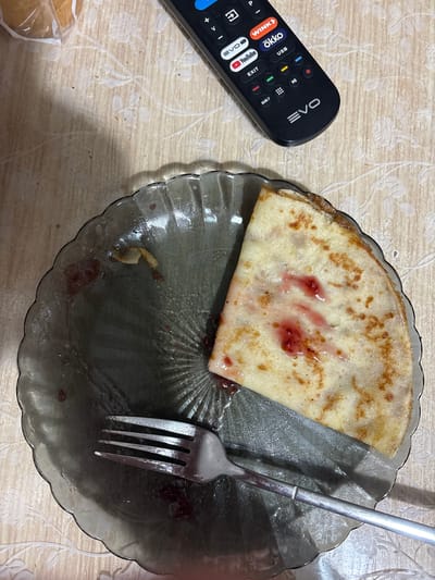 The image shows a plate with a partially eaten crepe topped with jam. A fork rests on the plate, which appears to be made of dark, translucent glass. A black TV remote control with various buttons and logos is visible in the upper right corner of the image, lying on a patterned tablecloth. The scene suggests a casual meal or snack. There is no information in the image to determine the location within Kstovo, Russia.