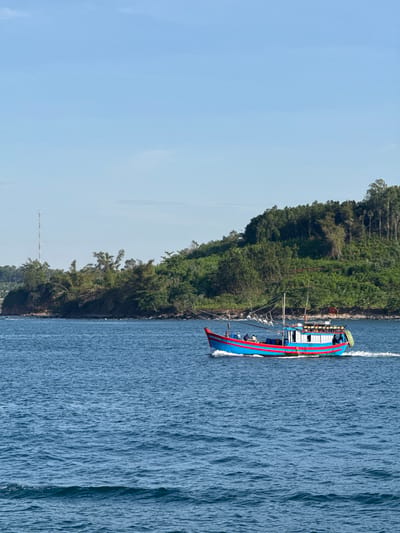A bright blue and red fishing boat is sailing on a clear blue sea. The boat has a white cabin with windows and is carrying several people on deck. In the background, a lush green, tree-covered island rises from the water. A tall communication tower is visible on the island. The sky is a clear, pale blue with a few wispy clouds. The water is calm, with small ripples on the surface. The scene appears to be a coastal area in An Hải, Vietnam, likely during the daytime.