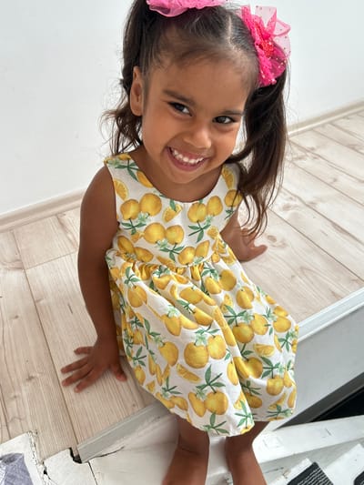 A young girl with dark hair, styled in pigtails with pink bows, smiles broadly while lying on a light-colored wooden floor. She is wearing a yellow dress with a pattern of lemons and green leaves. Her eyes are dark and wide, and her mouth is open, revealing her teeth. Her left arm is bent, with her hand resting on the floor. The background shows a white wall and part of a white structure that appears to be a piece of furniture or a doorway. The lighting suggests it is daytime. There is no visible text in the image. The setting is indoors, likely a home. The girl appears happy and playful.