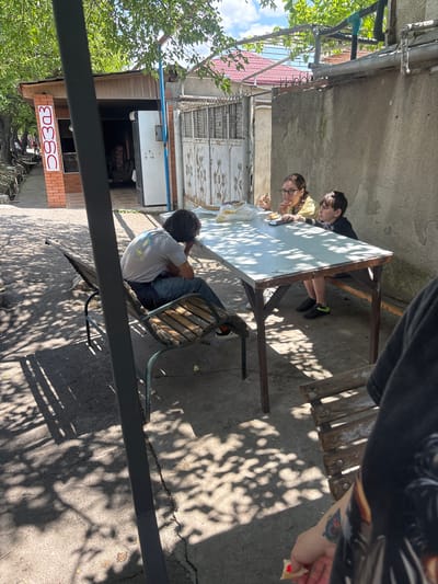 In Badiauri, Georgia, a group of people are gathered around a table outdoors, with a focus on a young woman and a boy eating. The woman wears glasses and a yellow shirt, while the boy has dark hair and a black shirt. They are seated at a light-colored table with wooden legs and a darker wooden frame. A person in the foreground is partially visible, holding a piece of food. To the left, a man with dark hair and a grey t-shirt is seated on a wooden bench, looking away from the camera. The scene is set in a paved area with trees overhead, casting dappled shadows on the ground. In the background, a brick building with a vertical sign and a white refrigerator is visible, along with a metal gate and a house with a red tiled roof. The overall environment suggests a casual outdoor gathering or meal.