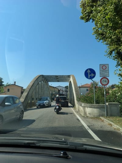 This is a photograph taken from inside a vehicle, looking out at a road leading over a bridge. The sky is clear and blue. On the right side of the road, there are two signs. The top sign is a rectangular white sign with text and a red circle with a line through it. The bottom sign is a circular sign with a red border and a red line through a bicycle icon, indicating no bicycles allowed. A person on a scooter is riding on the road, crossing the bridge, and other vehicles are also present. Buildings and trees are visible in the background. The location is specified as San Stino di Livenza, Italy.