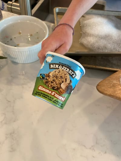 The image shows a person's hand holding a pint of Ben & Jerry's "Gimme S'more!" ice cream. The hand is wearing a purple bracelet. The ice cream pint is positioned in front of a white counter. Behind the pint is a partially visible baking sheet with soap bubbles. The scene is located in a kitchen in Rye, New York. The coordinates (latitude: 40.952, longitude: -73.708) suggest the location is a residential area in Rye, New York.