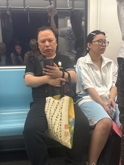 This media file shows a group of people on a public transport vehicle, likely a train or bus, in Xuhui District, China. The scene is dimly lit, suggesting it could be evening or a windowless interior.
In the foreground, a man is holding a smartphone, appearing to take a selfie or record a video. He is wearing a black t-shirt with a graphic on the left breast that partially reads "bonjour". He has a neutral expression. He is leaning against a bag that has Chinese calligraphy and imagery printed on it.
Behind the man, a woman wearing glasses and a white jacket is seated. She appears to be looking away from the camera, and her eyes are closed, suggesting she might be resting or sleeping. She is holding a smartphone in her right hand. She is wearing a light blue shirt underneath her jacket. Another person, partially visible, is sitting next to her. The overall mood seems relaxed, with people engaged in their own activities or resting during their commute. The blue walls of the vehicle are visible, along with reflections in the dark surfaces, hinting at other passengers or the interior of the transport. The visible text on the bag is Chinese calligraphy, but it is not fully legible.