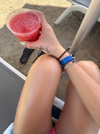 The image shows a person sitting on a beach chair with a red drink in their hand. They have multiple bracelets on their wrist, including a blue one with a black fastener and a thin brown one. A black tattoo is visible on the wrist. A pair of black flip-flops rests near the chair leg. The background includes a sandy area and part of the beach chair. The person's crossed legs are lightly tanned.