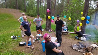 A group of seven people are gathered in a forest clearing, possibly for a picnic or an outdoor celebration. Two women and five men are present, with ages appearing to range from late teens to middle age. The setting is a natural environment with tall pine trees, green grass, and a dirt path. Several colorful balloons are tied to a tree, suggesting a festive occasion.
In the foreground, a woman with bright pink hair, wearing a black dress and sunglasses, is seated on the ground, looking towards the camera with a relaxed expression. To her left, a woman in a dark blue t-shirt and black shorts stands with her hand on her hip, making a peace sign with her other hand. Behind her, another woman in a light blue t-shirt and light-colored pants stands with her hands on her hips.
In the middle ground, a man wearing a gray t-shirt and shorts stands next to a small portable stove with a pot on top. Another man, in a black t-shirt and pants, stands to his right. Further back, a man in a camouflage jacket and green t-shirt sits on a wooden bench near a tent. A campfire is visible in the lower right corner of the image, with smoke rising.
Notable objects include camping gear like a tent and a portable stove, as well as a bench and a picnic table. The lighting suggests it is daytime, likely afternoon, with overcast or diffused sunlight. The overall mood seems casual and enjoyable, with some individuals smiling or posing. The t-shirt of the man in the middle ground has text that appears to read "FLORIDA".