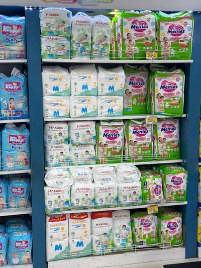 This is a retail display of baby diapers in a store. The shelves are stocked with various brands and sizes of diapers. Brands visible include "Baby Happy!", "Makuku", and "Merries". The diapers are packaged in brightly colored bags and boxes. Some packages have promotional tags indicating discounts. The environment appears to be an aisle in a supermarket or a dedicated baby product store. The presence of blue shelves and white price tags suggests a retail setting. The lighting is bright, illuminating the products clearly. The arrangement is organized, with products neatly stacked on shelves. The image captures a section of the store dedicated to baby care products.