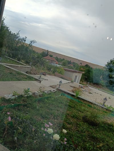 The image depicts an outdoor scene with a tilted perspective, showcasing a rural landscape with a house and surrounding vegetation. The sky is overcast with a light gray cloud cover. In the foreground, there is a grassy area with some pink and white flowers and a black decorative fence. Several white rectangular planters are visible, some containing small plants. A concrete pathway and retaining wall are also present, with garden lamps placed along them.
Further back, a small, gray, box-like structure with a sloped roof and a door is situated on a gravel-covered area. Beyond this structure, a traditional-style house with a red tiled roof is visible amongst trees. The background features rolling hills with cultivated fields and more trees. The overall impression is of a residential property in a rural setting, likely within the city of Suhache, Bulgaria, given the information provided. No people are visible in the image.