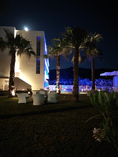 Captured at night in Gouves Municipal Unit, Greece, the scene features a well-lit building with a multi-story structure casting an artificial glow on the landscape. Several palm trees stand prominently, illuminated from the ground, adding to the exotic ambiance. In the foreground, white wicker chairs are neatly arranged on a lawn, suggesting an outdoor seating area. Further back, the faint glow from more distant lighting hints at additional structures or recreational areas beyond the immediate lawn. The darkness of the night sky contrasts sharply with the artificial lighting, creating a strong sense of depth and dimension within the frame.