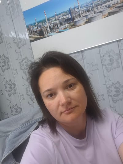 A mid-shot selfie shows a woman with shoulder-length, dark brown hair in a light purple t-shirt and a denim jacket draped over her shoulders. She is looking at the camera with a neutral expression. Behind her, a patterned wallpaper features a floral design. In the background, a large framed picture displays an industrial landscape with tall chimneys and storage tanks under a clear blue sky. The image appears to be taken indoors, possibly in a residential or office area of the Tecnimont camp in Russia.