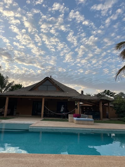 The image shows a single-story structure with a thatched roof, located next to a swimming pool. The building has a triangular window near the peak of the roof and several visible pillars supporting an overhanging roof that extends over a patio or deck. The pool has light blue water and a beige or light brown deck. Palm trees are visible in the foreground and background, suggesting a tropical environment. The sky has a dense scattering of cumulus clouds. The photograph appears to have been taken during the day, with a warm, indirect light. The location is likely Communauté rurale de Malicounda, Senegal, based on the geographical context.