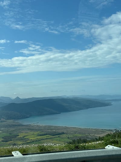 This image presents a panoramic landscape view overlooking Gökova Bay, near Akyaka, within the Ula district of Muğla Province, Turkey.

In the immediate foreground, a grey metal guardrail with visible bolts and white plastic end caps runs horizontally across the bottom of the frame, indicating the vantage point is from an elevated roadside or overlook. Sparse, dry green and brown vegetation grows on the rocky slope directly below the guardrail.

The middle ground features a relatively flat coastal plain stretching alongside the bay. This plain is segmented into numerous rectangular parcels of land, some distinctly green, suggesting agricultural fields, while others appear fallow or less cultivated. Towards the shoreline, a small cluster of white and light-colored structures, possibly buildings or a small settlement, is visible at the edge of the water.

The central feature of the image is a large body of calm, turquoise-blue water, identified as Gökova Bay. The bay's waters reflect the clear sky and are bordered by a curving coastline.

In the background and extending into the mid-distance, a series of densely forested hills and mountains rise from the water's edge, forming the natural perimeter of the bay. These mountains exhibit varying shades of deep green and blue due to distance and atmospheric haze, with distant peaks fading into lighter blues along the horizon.

The sky occupies the upper portion of the image, displaying a bright blue color with scattered white, layered clouds, indicative of clear weather conditions. No individuals, animals, or specific actions are discernible within the frame.