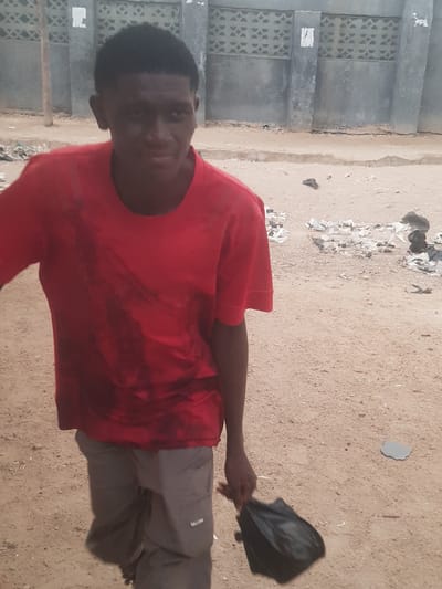 A man wearing a red shirt and gray pants stands on a sandy surface. He holds a black bag in his right hand. To the left of the man is a concrete building with decorative openings. Scattered trash is on the ground. The location is Dutsinma A, Nigeria.