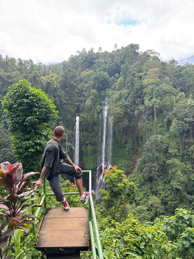 A man balances on a platform with railings, overlooking a lush, green forest landscape. Multiple waterfalls cascade down the verdant hillside. The man wears dark clothing and red and white sneakers. He seems to be stretching or performing a yoga pose, gripping the railing with one hand. The platform is wooden and surrounded by dense foliage. Brightly colored plants are visible in the foreground. The sky is cloudy.