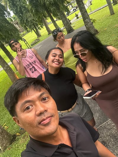 The image depicts a group of five individuals in Bedulu, Indonesia, captured in an outdoor selfie. In the immediate foreground, an adult male, likely of Asian descent, with short dark hair and a black polo shirt, looks directly at the camera with a neutral expression. Behind him and slightly to his right, an adult female, with short dark hair, a nose ring, and hoop earrings, smiles broadly at the camera. She is wearing a black high-neck top and striped grey trousers. An orange strap is visible over her right shoulder. To her right, another adult female with long dark hair, wearing oval-shaped sunglasses and a brown spaghetti-strap dress, holds a white smartphone horizontally in her left hand, looking downwards. Further back, positioned behind and slightly to the left of the smiling female, an adult female with dark hair pulled back and black sunglasses on her head, wears a light green strapless top, her gaze directed away from the camera. In the background to the left, an adult male with a beard and a backward black baseball cap wears a pink sleeveless basketball jersey featuring Chinese characters and the number "22." He holds a lit cigarette to his mouth with his right hand. The setting is a paved dark-colored pathway bordered by lush green grass and numerous tall palm trees. In the distant background, stone structures, possibly temple architecture, are partially visible, along with a white golf cart on a separate paved area and small stone decorative elements on the grass. The lighting suggests natural daylight.