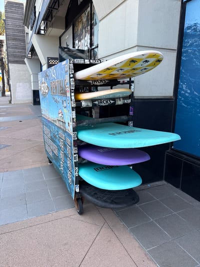 This image, rotated, displays a colorful array of surfboards vertically held in an outdoor rack, likely associated with a surf shop or rental in Huntington Beach. Six surfboards are visible. On the far left, a white surfboard features a distinctive yellow diamond pattern, each diamond containing a black palm tree silhouette. Next to it is a light, possibly beige, surfboard. These are followed by three vibrant boards: a light turquoise, a purple, and another light turquoise, all of which display "ALLON" printed vertically. The final surfboard on the right is dark grey or black, possibly covered.
The metal surfboard rack is extensively covered in an assortment of stickers and graffiti. Visible text from these decals includes "JACKS SURFBOARDS" (appearing multiple times), "NEWREKIS", "EMON", "HEU ISHU", "SONER", "SCV", "TLF", "3XK", "BOR", "CHUR", "ANE", "LEKO", "BA", "NEZE", and "SIDH." The lower front panel of the rack is a large blue board depicting a stylized ocean scene with waves, over which white text and additional graffiti are layered, making some parts illegible, though "REMIND" can be partially seen.
In the background, a building facade with windows and dark framing is visible. Behind the surfboards, there's a dark blue wall or display panel featuring a textured design resembling ocean water. The ground is paved with light-colored square tiles. The scene is brightly illuminated, indicating daytime conditions. The surfboards and the overall aesthetic strongly suggest a coastal, surf-centric environment.
