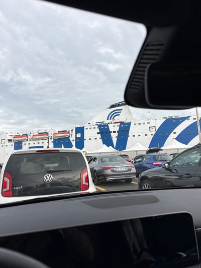 The image depicts a view from inside a car, likely waiting in line on a ferry in Civitavecchia, Italy. Visible through the windshield is a large ferry ship, predominantly white with blue stripes, docked beside a queue of cars. Several cars are visible in front and to the side. The sky is overcast. The interior of the car from which the photo is taken is partially visible on the left and right edges of the frame. A dashboard display is on the right.