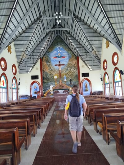 The image shows a person walking down the aisle of a church in K'Rèn, Vietnam. The church interior features a high, vaulted ceiling with exposed metal beams and corrugated metal roofing. Rows of wooden pews line both sides of the aisle, facing a large mural at the far end depicting a crucifixion scene with Jesus Christ on the cross, God the Father above, and a depiction of the Virgin Mary. Stained-glass windows are visible on the side walls, and statues of religious figures are placed in niches. The floor is polished and reflective. The person walking away from the camera is wearing a blue t-shirt, grey shorts, a backpack, and sandals.