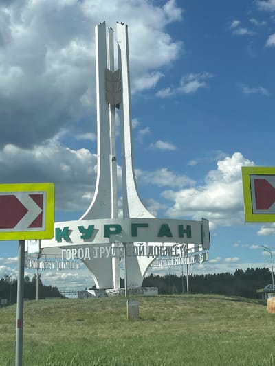 The image shows a monument with the inscription "КУРГАН" (Kurgan) in large green letters, followed by smaller white letters that read "ГОРОД ТРУДОМ И ДОБЛЕСТЬЮ" (City of Labor and Valor). This monument is located on a grassy hill, likely at the entrance to the city of Kurgan, Russia. There are directional signs on either side of the monument, one showing a rightward arrow and the other a leftward arrow, both with red and white markings within a yellow border. In the background, a line of trees forms the horizon, and beyond that, a bridge-like structure is visible. The sky is blue with scattered white and dark clouds. The overall scene depicts a civic monument marking the city's boundary.