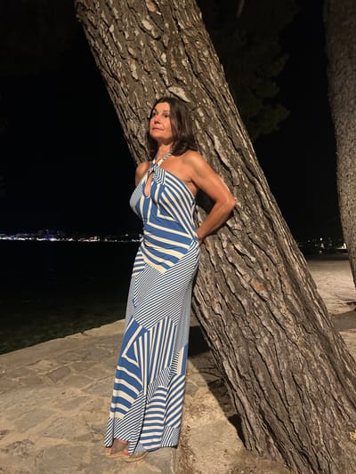 A woman leans against a large tree trunk at night in Port de Pollença, Spain. She is wearing a blue and white striped halter dress and espadrille sandals. The background shows a dark body of water with a distant shoreline illuminated by city lights. The ground is paved with stone.