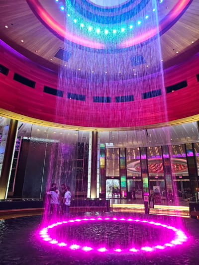 The photograph depicts an interior space within the Seminole Hard Rock Hotel & Casino Hollywood, located in Hollywood Seminole Reservation, United States of America.
A major element is a multi-colored fountain featuring strands of water illuminated in hues ranging from blue to pink and purple. The fountain has a circular design and the cascading water forms a visual screen.
In the foreground, two individuals are standing on a platform. They are positioned near the fountain, appearing to be within its range. Their attire includes white tops. The angle suggests they are likely observing or interacting with the water feature.
The background consists of architectural elements of the interior of the casino and hotel. These include reflective surfaces, a mezzanine with glass barriers, and design motifs typical of the Hard Rock brand. The walls and ceilings incorporate panels in shades of red, brown, and gold. A ring of lights are along the side of the building. The scene is well-lit with colored lights, highlighting the fountain.