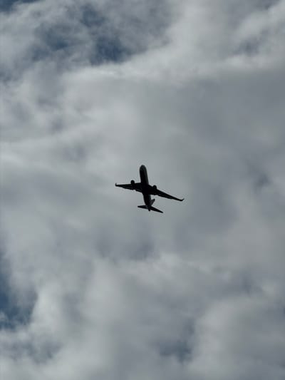 An airplane is silhouetted against a cloudy sky. The airplane is captured from below, appearing to be climbing, with its landing gear extended. The clouds are a mix of white and gray, obscuring parts of the sky. The image does not provide any contextual information to determine its location within Kostelec u Křížků, Czechia.