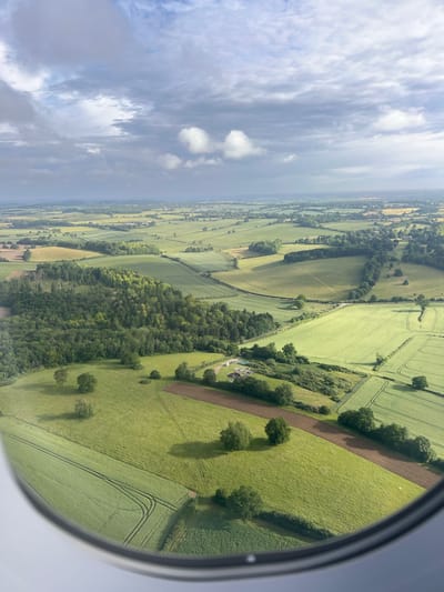 An aerial view from an airplane window shows a patchwork of green fields and trees under a cloudy sky. The landscape is rural, with distinct fields delineated by hedgerows and tree lines. Several agricultural fields are visible, some with tire tracks from machinery. A cluster of trees forms a dense woodland in the mid-ground. In the distance, more fields and scattered buildings suggest a rural settlement. The foreground is dominated by a section of green field with tire tracks and scattered trees, framed by the circular edge of the airplane window. The location is the rural area of North Hertfordshire, United Kingdom.
