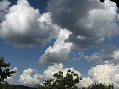 The image shows a view in Drianovets, Bulgaria, with a predominantly cloudy sky. The sky is partially blue, but large, puffy white and gray clouds dominate the frame. The clouds vary in density and color, with some appearing lighter and brighter than others. In the foreground, there are trees with green foliage, suggesting a natural environment. The overall scene depicts a typical day in Drianovets with a combination of sunshine and potential for rain.