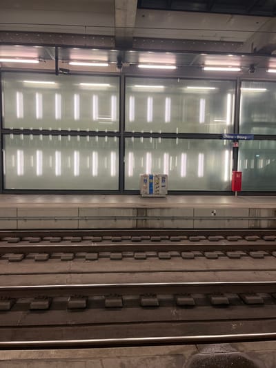 The image displays an empty, modern train or metro station platform and tracks, likely an underground or covered facility. No people are visible in the scene.
The composition is divided into two main sections by a clear glass partition that runs horizontally across the middle of the frame. To the left of this partition is the station platform area, featuring a concrete wall on the far left and multiple vertical, linear fluorescent light fixtures. These lights cast prominent, rectangular reflections onto the glass partition, creating a pattern of bright horizontal stripes.
To the right of the glass partition are the train tracks. Three sets of parallel rails are clearly discernible, running across the frame from left to right. The tracks are laid on a concrete bed with regularly spaced rectangular elements, which appear to be track fastenings or sleepers. The area between and around the tracks consists of concrete, with some darker, possibly wet patches on the far right edge.
Key details include a blue rectangular sign mounted on the glass partition, bearing the white text "Chêne-Bourg". Below this sign is a small red box, likely an emergency or signaling device, with indistinct white symbols. Further along the glass partition, a small utility or control panel with various labels and indications is attached.
The environment is characterized by its functional, contemporary design, utilizing materials such as concrete and glass, and relying on artificial lighting. There is no activity taking place, and the station appears to be quiet and unoccupied.