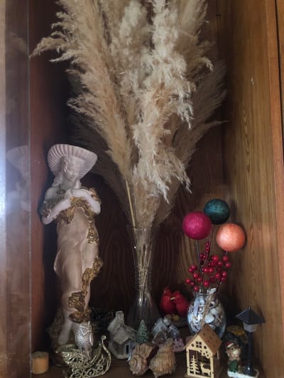 This is a photograph taken indoors, focusing on a shelf filled with various decorative items. The shelf appears to be made of wood, with a warm, brown tone and visible grain. Dominating the center of the shelf is a large bouquet of dried, feathery reeds in a clear glass vase. To the left of the reeds is a light-colored statue of a woman wearing a wide-brimmed hat, holding a bird or dove. The statue is adorned with gold-colored vine-like details. Below the statue, a small, glittery sleigh with an owl figurine is visible, along with a small, orange candle.

To the right of the reeds, a collection of decorative spheres on thin stems is arranged, with colors including dark teal, pink, and orange. Below these spheres, a glass vase filled with seashells holds a branch with red berries. Further to the right and towards the front, a miniature wooden house with cutouts of trees and a deer is displayed, next to a small figurine of a deer wearing a Santa hat, and a miniature black lamppost. Other small decorative houses, seashells, and figurines are scattered across the shelf. The lighting suggests the photograph was taken in natural light or with soft interior lighting, casting subtle shadows. The context of Chiatura, Georgia, is not visually discernible from the image itself; it is an external piece of information.