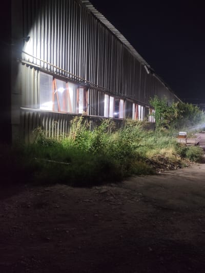 The image shows the side of a corrugated metal building at night, with a series of windows illuminated from within. A bright outdoor light illuminates the left side of the building. Tall grass and bushes grow in front of the building. In the background, there are trees and a small trailer with what appears to be a grill on it. The ground in front of the building is dirt and gravel. The scene is located in the city of Dangir, Uzbekistan.