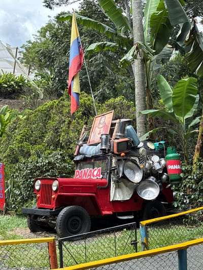 This image captures a static outdoor scene in Quimbaya, Colombia. A red, vintage-style Willys Jeep, marked with "PANACA" on its windshield, is positioned centrally. The vehicle is heavily laden with a diverse assortment of household items: a framed religious painting (depicting a figure resembling Jesus Christ), an old television set, and numerous metallic pots, pans, and other kitchenware stacked on its roof and hanging from its sides. A green propane gas cylinder, also bearing "PANACA," is secured to the vehicle's rear passenger side.

Behind the jeep, a Colombian flag, displaying vertical yellow, blue, and red stripes, flies from a pole. The surrounding environment consists of dense, lush green vegetation, including large trees with broad leaves (consistent with banana plants) and various bushes. In the distant left background, a white, net-like structure, typical of agricultural cultivation, is partially visible. A low, two-tiered fence, featuring yellow top rails and a chain-link mesh, occupies the immediate foreground, separating the viewer from the jeep. The ground directly beneath the vehicle is exposed earth with minimal vegetation. The sky overhead is overcast. No people are present, and no actions or interactions are depicted.