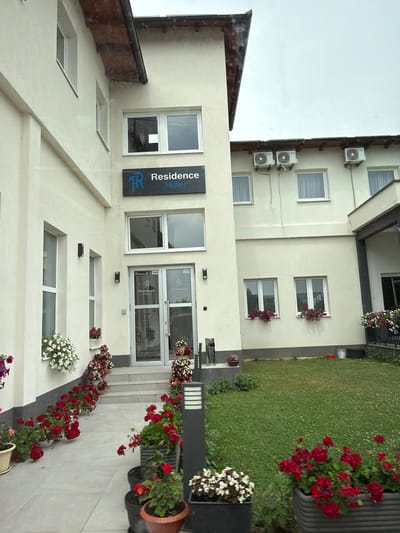 The image shows the exterior of a hotel named "Residence Hotel" located in Nević Polje, Bosnia and Herzegovina. The hotel is a light-colored, multi-story building with several windows. In front of the building, there is a paved walkway leading to the entrance, which features double glass doors. The walkway and the building's facade are adorned with numerous potted plants and flower arrangements, predominantly featuring red and white flowers. A well-maintained green lawn is visible to the right of the building, and several outdoor air conditioning units are mounted on the upper exterior wall. The sky is overcast.