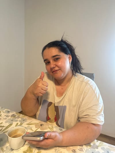 A woman with dark hair pulled back in a ponytail, wearing a light-colored t-shirt with a graphic design, is seated at a table in what appears to be a dining area, with a floral-patterned tablecloth. She is looking towards the camera and giving a thumbs-up with her right hand, while holding a smartphone in her left hand. Next to her phone on the table is a white coffee cup with a dark liquid inside. The background shows a plain, light-colored wall and a part of a grey chair. The lighting suggests an indoor setting. Based on the provided information, this scene is located in the city of Mainburg, Germany.