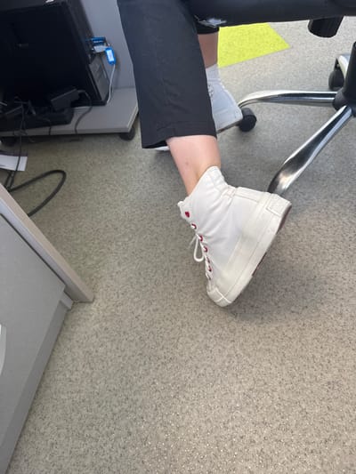 A person is sitting in an office chair with their feet up, resting on a desk. They are wearing white high-top Converse sneakers with red laces and red heart eyelets. The person's legs are covered by black pants. In the background, a computer tower and other office equipment are visible. The flooring is a speckled gray carpet. The scene is indoors, likely in an office setting. There is no information available to determine the location in the city of Suceava, Romania.