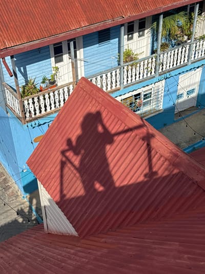 An elevated view reveals a building with blue and white walls, ornate white balconies, and windows with decorative grilles. Lush greenery adorns the balconies. Adjacent buildings are topped with red corrugated metal roofs. A distinct shadow, possibly cast by a chair or similar object, is projected onto the red roof. The scene is brightly lit.