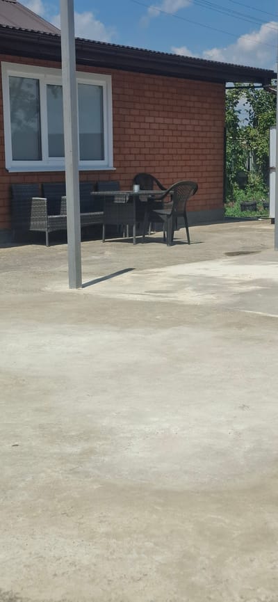 The image shows a patio area in front of a brick building, possibly in Kabekhabl, Russia. In the background, there is a building with a brown roof and a white-framed window. To the left of the building, there is outdoor furniture, including what appears to be a wicker sectional sofa and a table with chairs. The ground is paved with concrete. The overall impression is of an outdoor seating area, possibly for a cafe or residential property.
