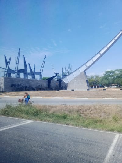 The image displays a daytime view of the Campo Carabobo Monument in Campo Carabobo, Venezuela, under a clear blue sky. In the background, the monument features a large, light-colored concrete and metallic structure rising diagonally upwards towards the right from a substantial concrete base. To its left and behind it, multiple dark, angular, 'V'-shaped metal sculptures are arranged on an elevated concrete platform. Below these sculptures, a dark bas-relief frieze depicting historical figures, some on horseback, is visible along a lower concrete wall.

In the mid-ground, a male individual is riding a dark-colored bicycle from left to right on a paved road. He is wearing a blue shirt, blue trousers, and a light-colored straw hat. A brown box or crate is attached to the handlebars of his bicycle. The immediate foreground consists of a paved asphalt road with visible white lane markings. Between the road and the monument, a strip of ground is covered with dry, sparse grass interspersed with patches of green vegetation. Deciduous trees are visible in the far background on the right side of the frame.