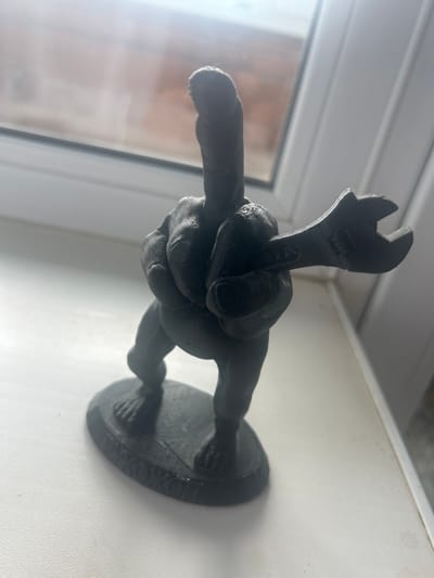A dark, 3D printed figurine of a person holding a wrench stands on a white surface, possibly a windowsill, in Kokhma, Russia. The figurine is black and appears to be made of plastic. It has a somewhat abstract design, with visible layers from the printing process. The person's stance is dynamic, and they are grasping a wrench in their right hand. The background is blurred, showing what appears to be a window with some light coming through, and possibly a brick wall outside. The overall impression is of a decorative item placed on a flat surface.