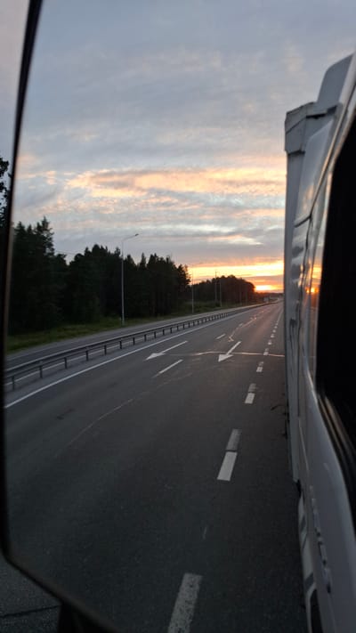 The image depicts a view from a vehicle traveling on a highway during sunset. The highway has multiple lanes marked with white lines and arrows, and a guardrail separates it from a treeline on the left. The sky is cloudy with streaks of orange and yellow from the setting sun. A portion of a white truck is visible on the right side of the frame. The location is identified as the city of Богандинский, Russia.