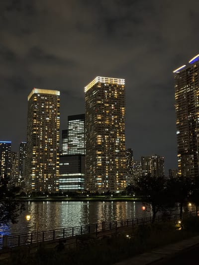 This is a nighttime photograph of a waterfront cityscape in Koto, Japan. The scene is dominated by numerous illuminated high-rise buildings, their windows glowing with warm yellow light against the dark sky. The buildings vary in height and architectural style, with some featuring sharp, modern designs and others appearing more rounded.

In the foreground, a dark, calm body of water reflects the lights from the buildings and the sky. A railing-lined walkway or promenade runs along the edge of the water, with scattered streetlights casting pools of illumination. Dark trees and foliage line the embankment, partially obscuring the lower portions of the buildings. The sky is overcast with dark clouds, contributing to the nighttime atmosphere. No people are visible in the frame.