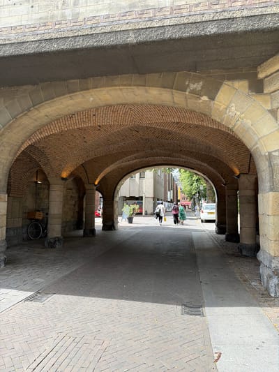 This is a photograph taken from beneath a brick-arched overpass, likely a bridge, on a sunny day in Roermond, Netherlands. The overpass is supported by thick stone pillars, with visible graffiti on some of them. The ground is paved with brick in a herringbone pattern, with a drain grate visible in the foreground. On the left side, under the overpass, a bicycle with a wooden crate on the back is parked next to a stone wall. The perspective looks out towards a street where a white car is parked on the right. A few people are walking away from the camera down the street, with buildings and trees visible in the background. The overall impression is of an urban environment with historical architectural elements.